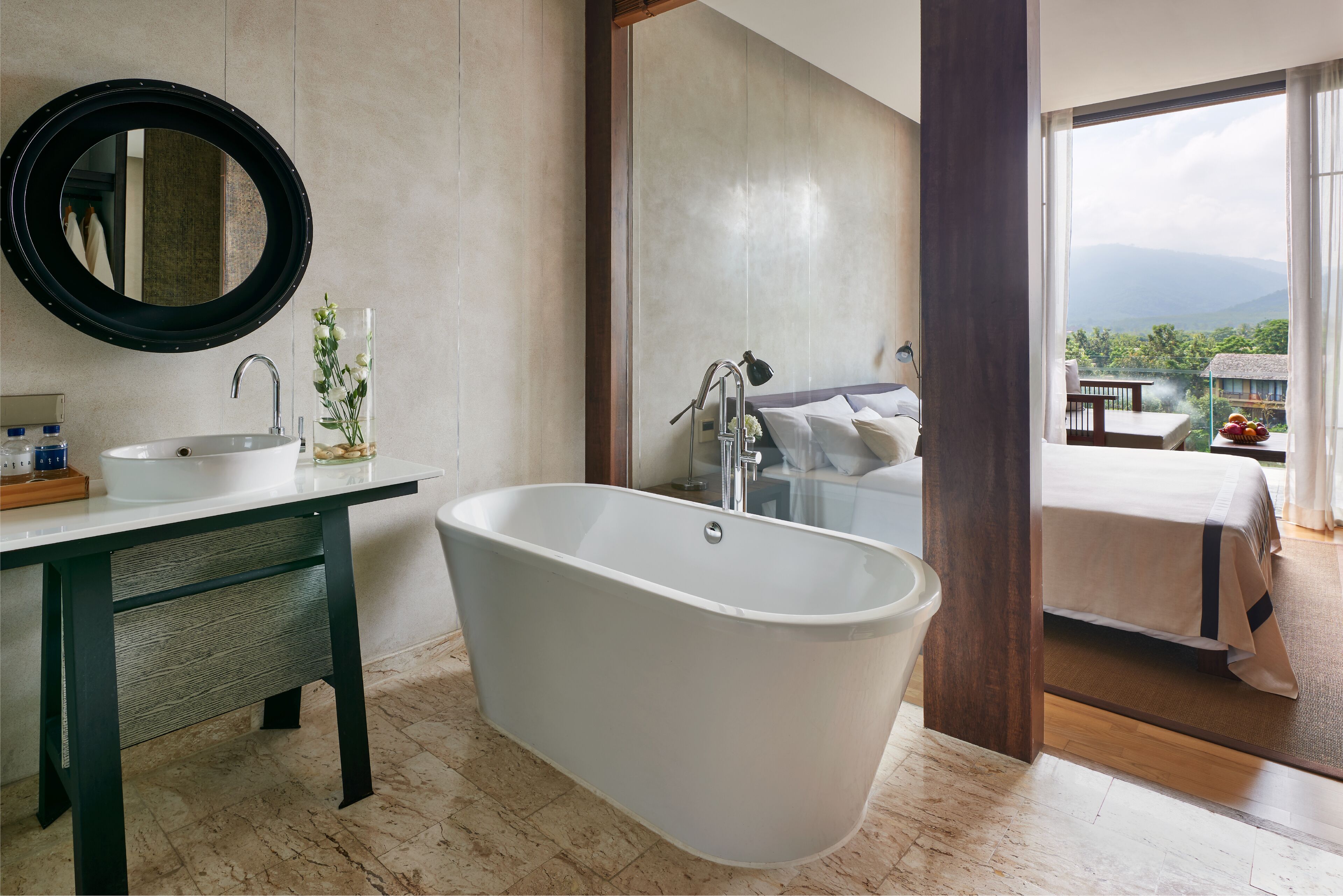 Un baño moderno con una bañera independiente, un espejo redondo y un lavamanos. Un gran ventanal ofrece vistas a un balcón con una cama y una mesa.