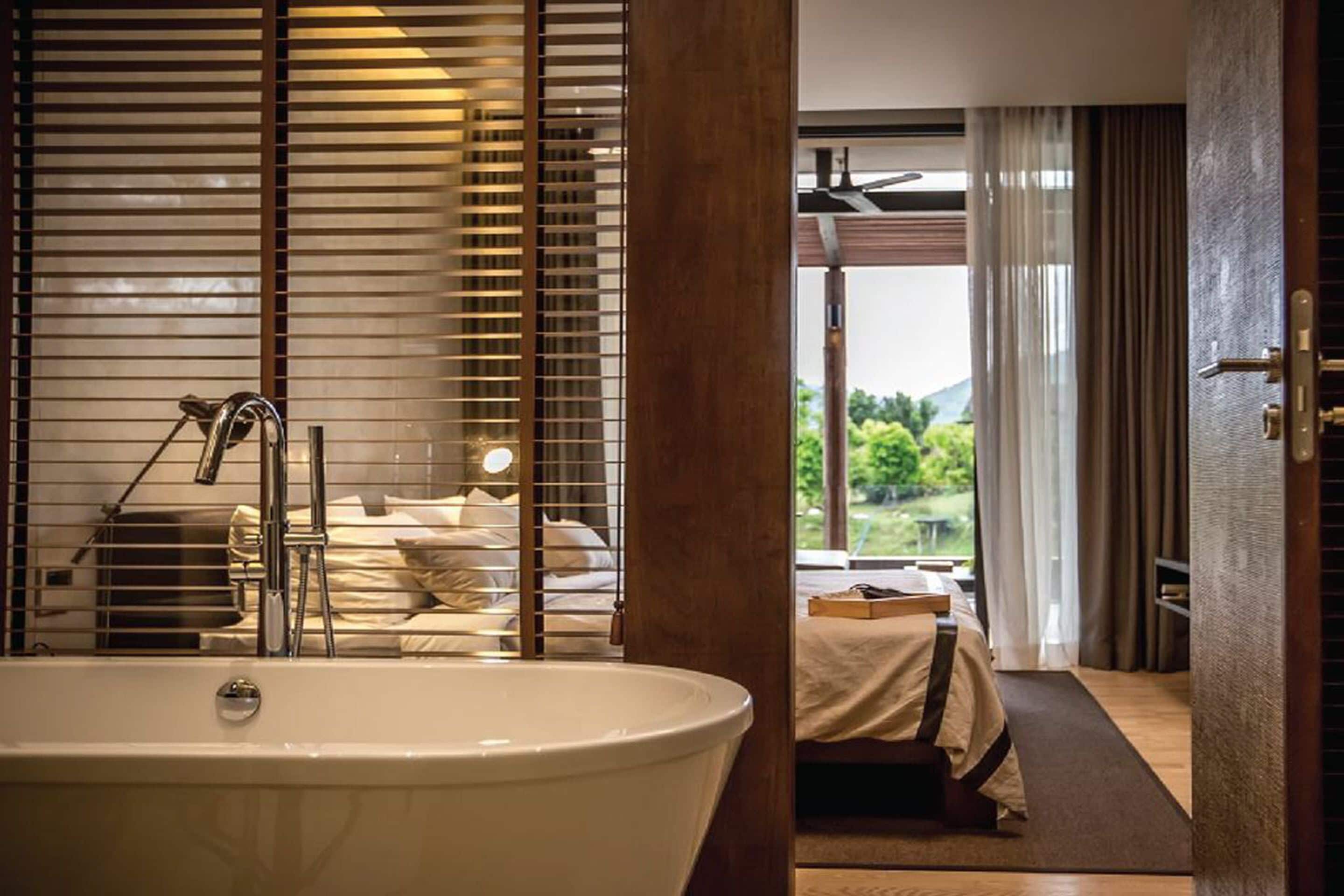 A modern bathroom with a large bathtub, a shower area, and a view of a bedroom with a bed and a window.