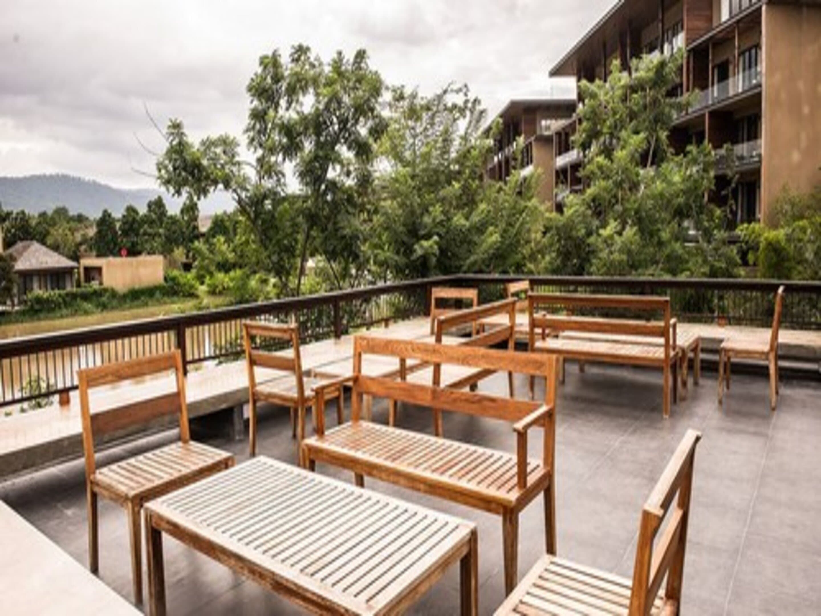 An outdoor seating area with wooden tables and benches, surrounded by greenery and a view of a building in the background.