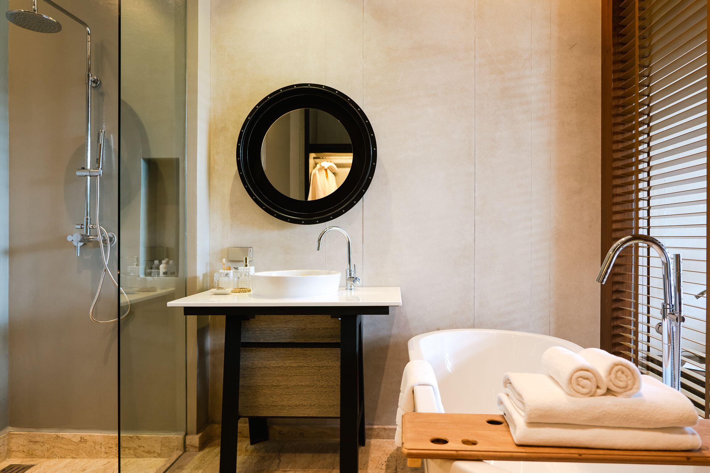 A modern bathroom with a glass-enclosed shower, a round mirror, a white sink, and a bathtub with rolled towels.