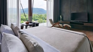 A modern hotel room with a large bed, a TV, a dining area with a table and chairs, and a view of the mountains. - atta Lakeside Resort Suite (Pak Chong)