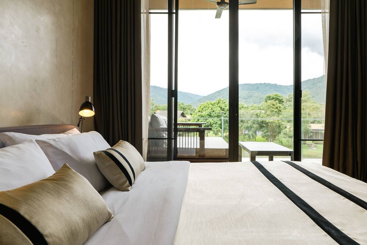 A hotel room with a large bed, a nightstand with a lamp, and a balcony with a table and chairs.