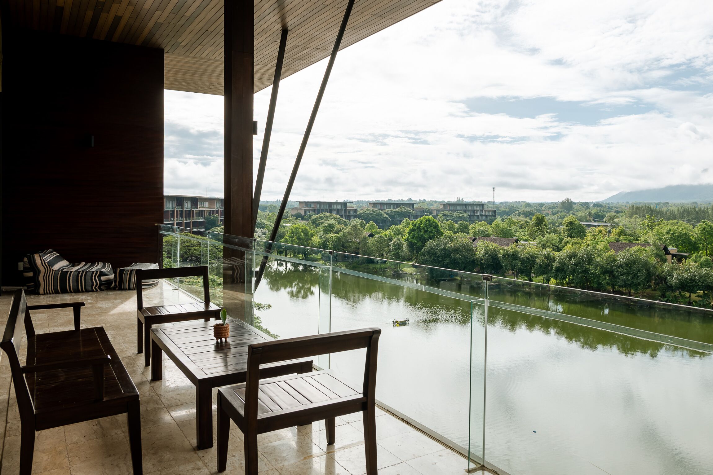 A balcony with a view of a river and greenery, wooden furniture, and a glass railing.