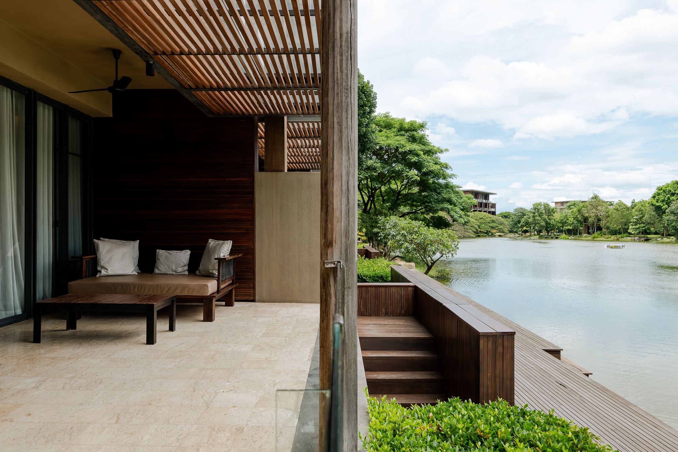 A modern outdoor seating area with a wooden bench and cushions, overlooking a calm body of water and lush greenery.