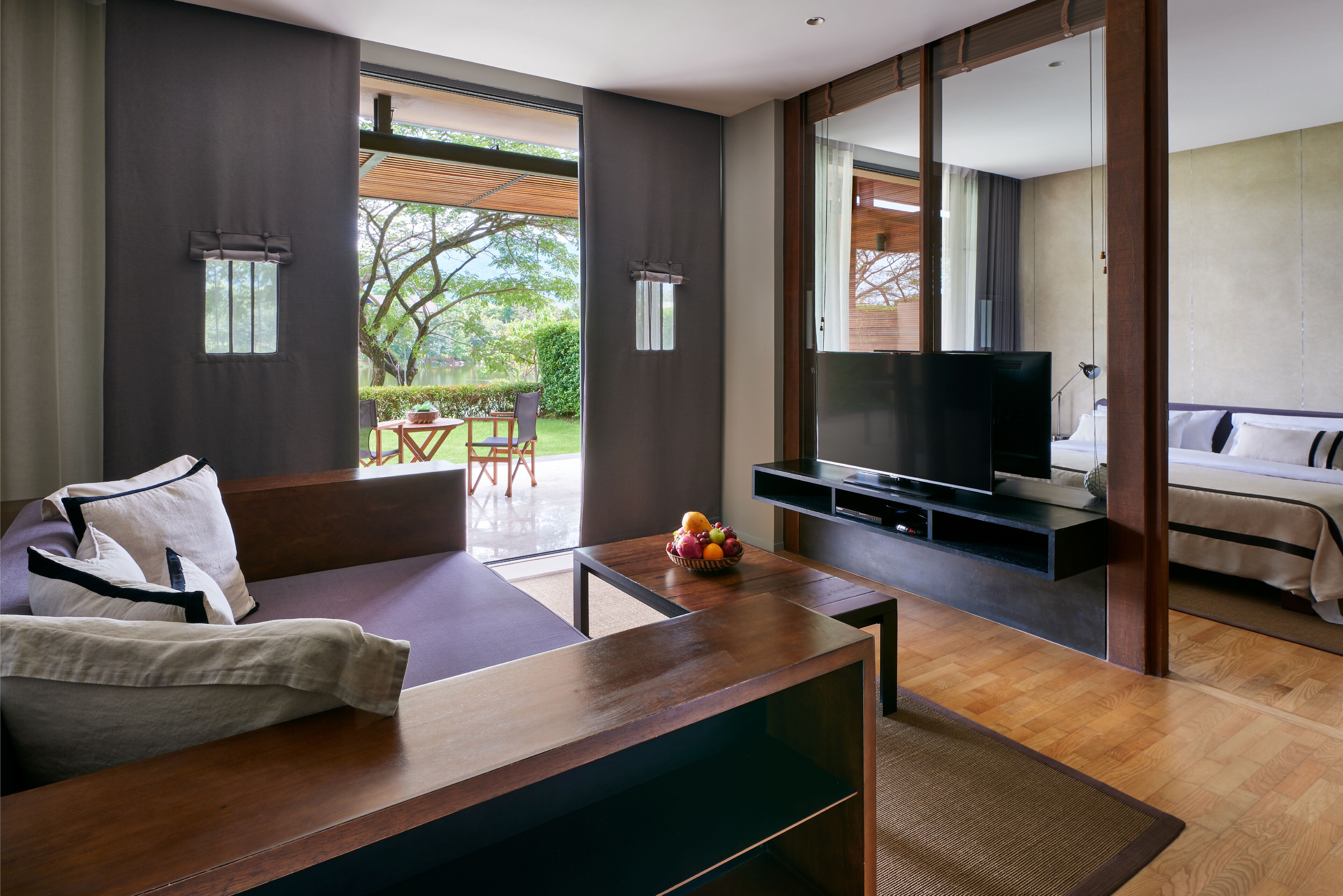 A modern living room with a sofa, a coffee table with fruit, a TV, and a balcony with a view of trees.