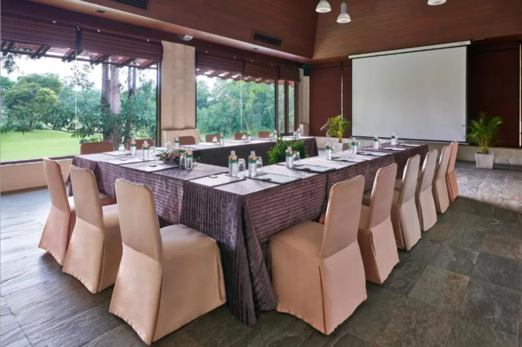 A conference room with a long table, chairs, a projector screen, and a view of greenery outside.