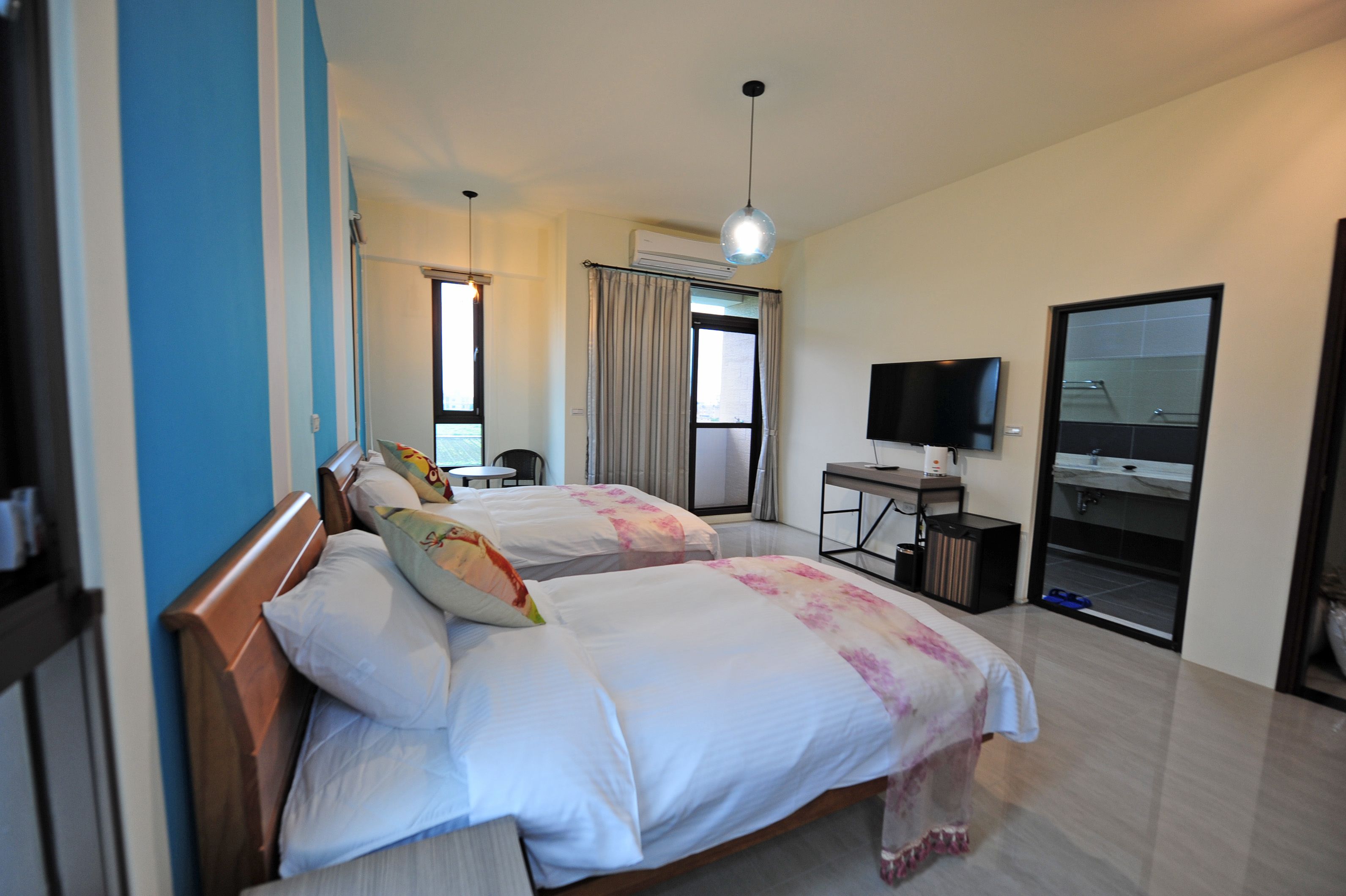 A hotel room with two beds, a desk with a flat-screen TV, an air conditioning unit, and a large window with curtains.