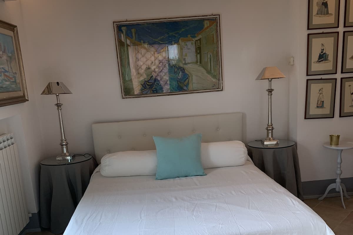 A neatly arranged bedroom with a large bed, a headboard, a bedside table with a lamp, a small side table, and a framed painting on the wall.