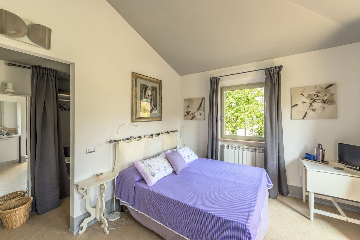 A bedroom with a bed, a desk, a window with curtains, and a view of greenery outside.