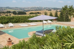 A swimming pool with lounge chairs and an umbrella, surrounded by greenery and a view of rolling hills. - Relais Il Margarito (Montefano)