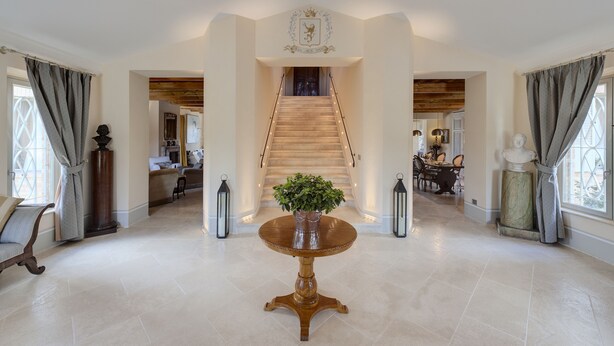 A spacious foyer with a grand staircase, a round wooden table with a plant, and a coat of arms on the wall. - Relais Il Margarito (Montefano)