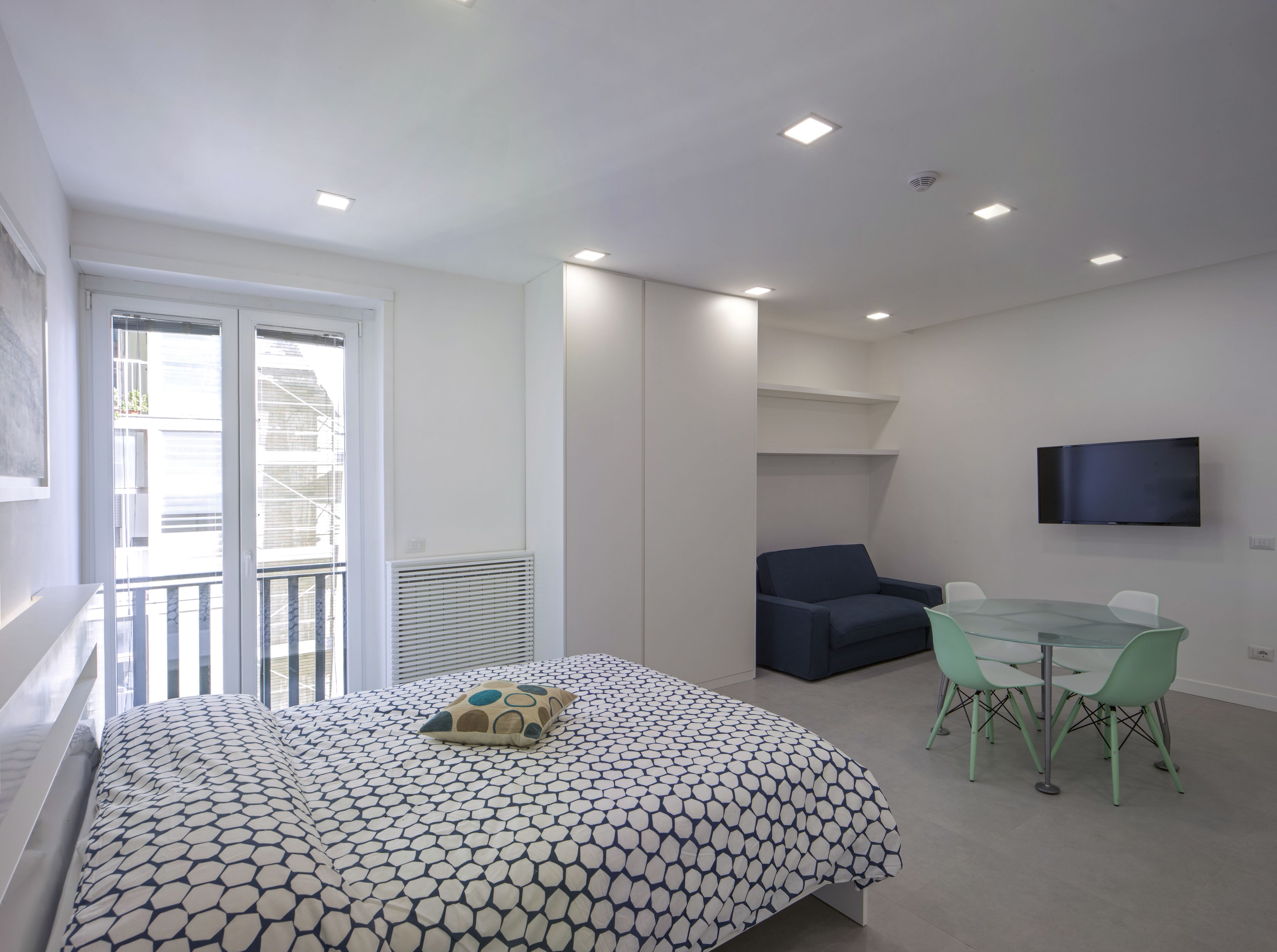 A modern bedroom with a bed, a seating area with a table and chairs, a TV mounted on the wall, and a balcony with a view of the outside.