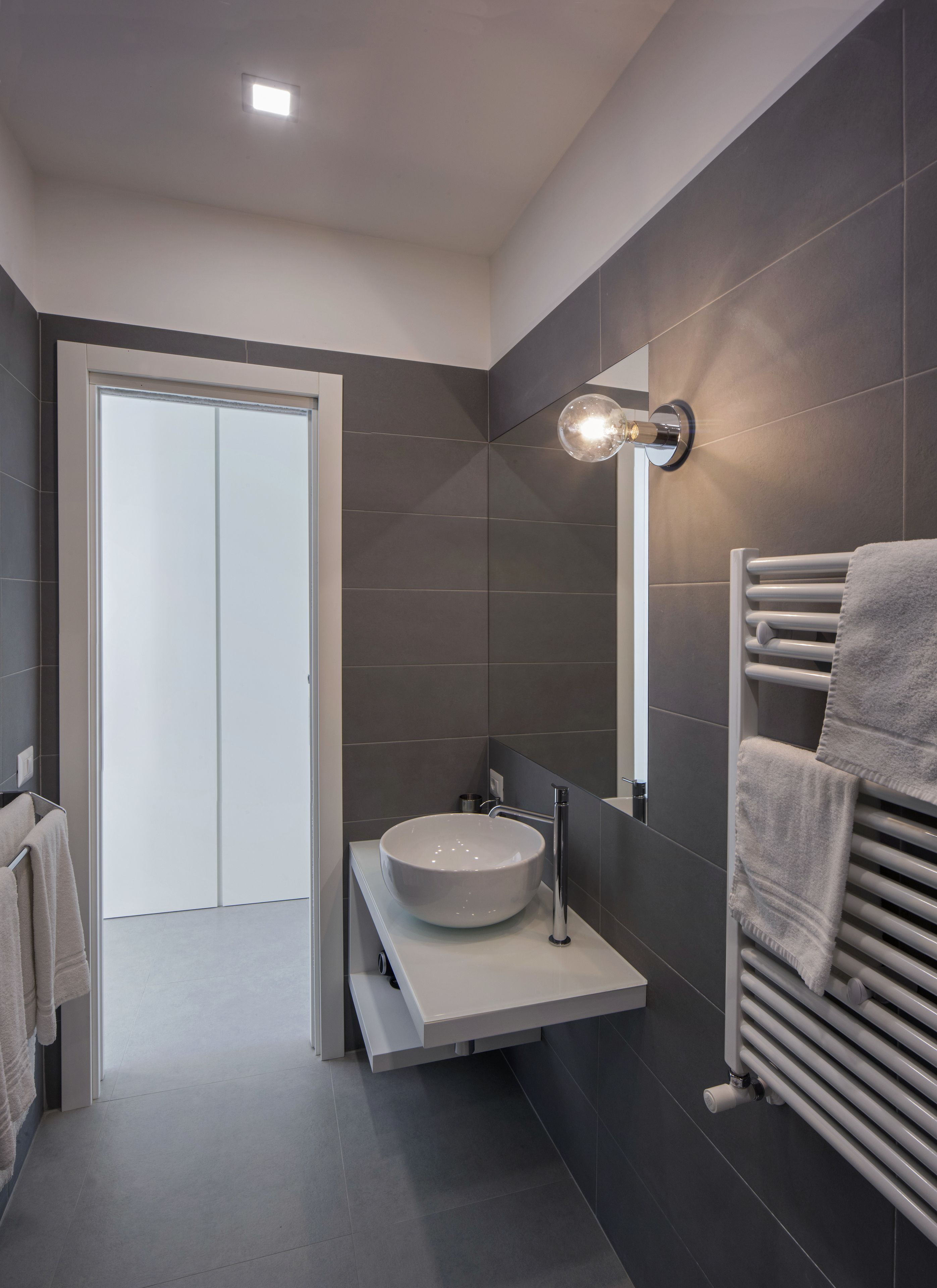 Ein modernes Badezimmer mit einem weißen Waschbecken, dunkel gefliesten Wänden, einem Handtuchhalter und einer Tür zu einem anderen Raum.