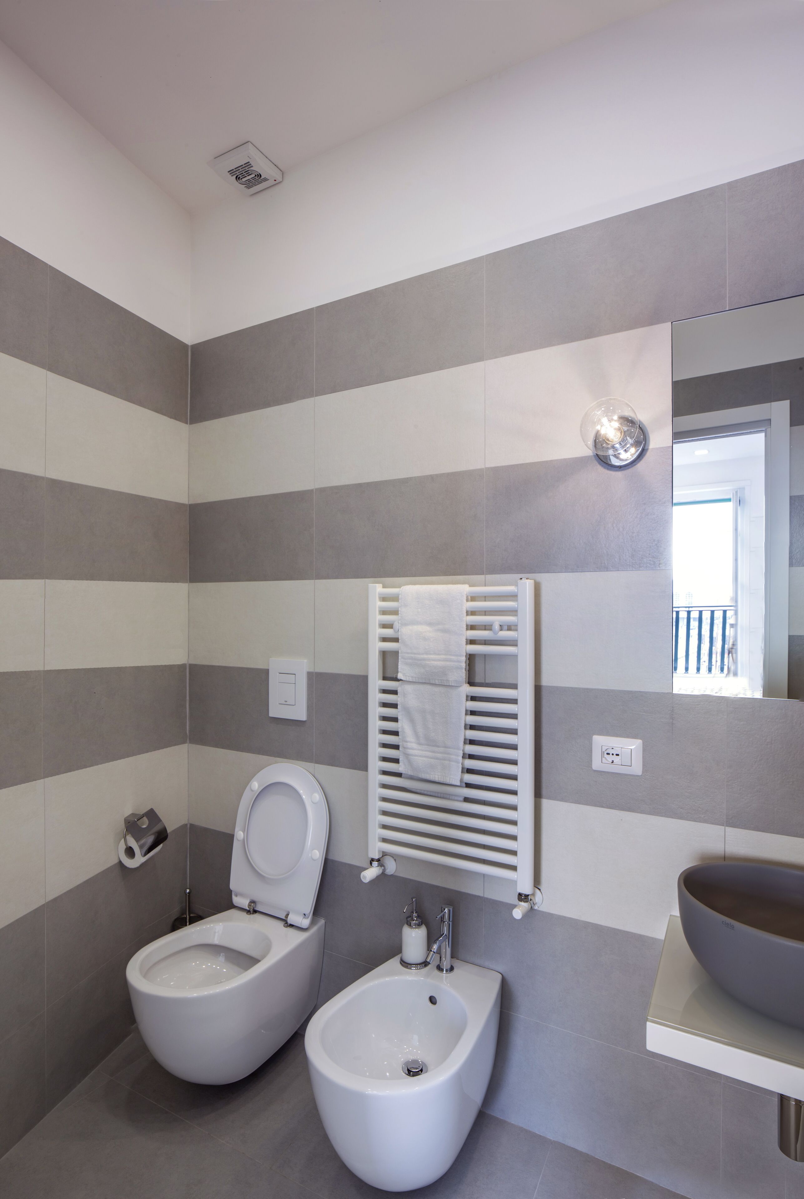 Een moderne badkamer met toilet, bidet en wastafel, met gestreepte betegelde wanden en een grote spiegel.