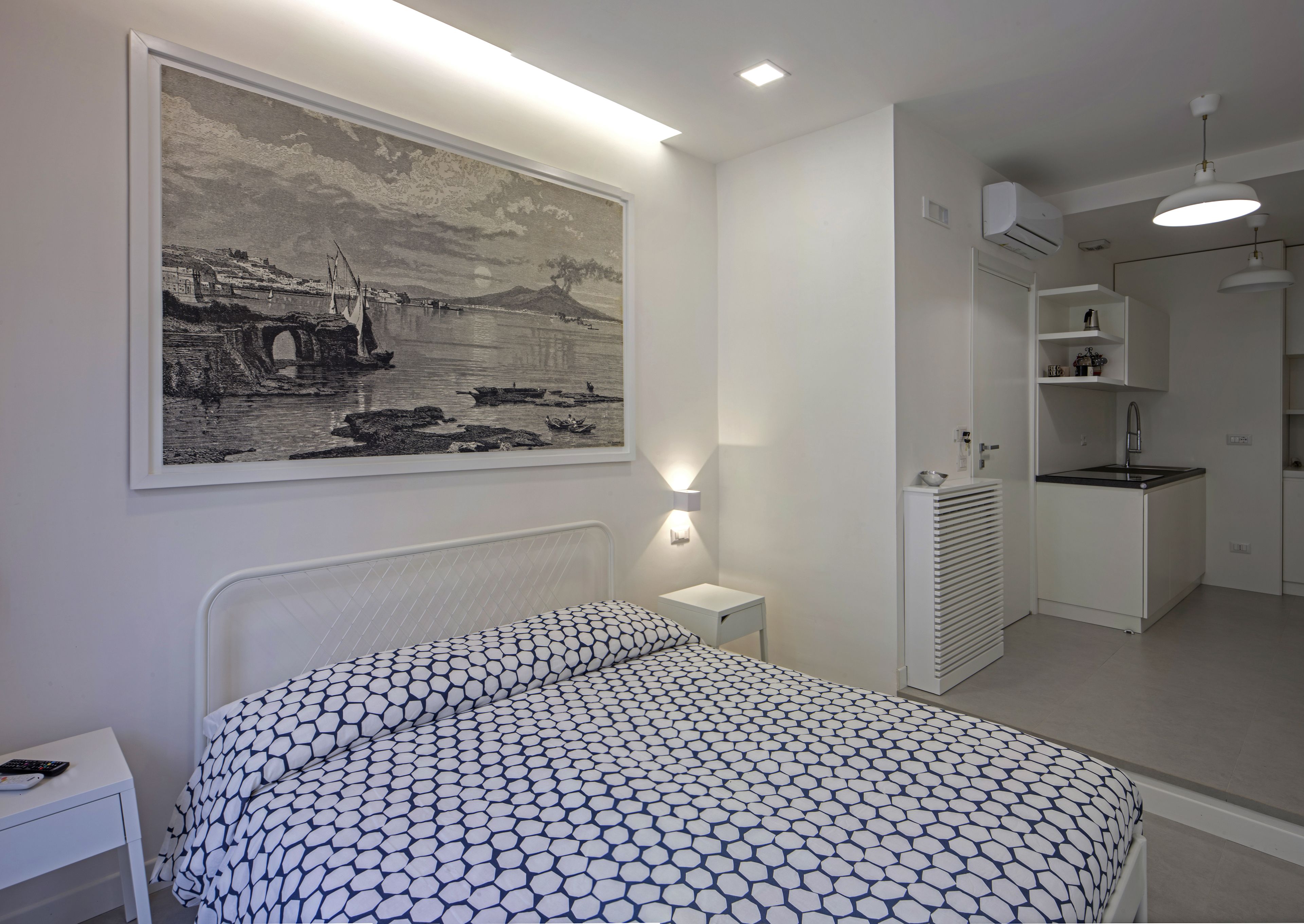 A bedroom with a bed, a nightstand, a wall-mounted lamp, a large framed picture, and a kitchenette area.