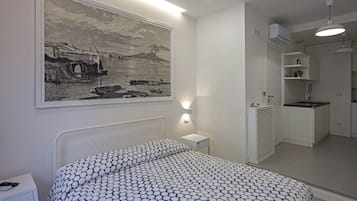 A bedroom with a bed, a nightstand, a wall-mounted lamp, a large framed picture, and a kitchenette area.