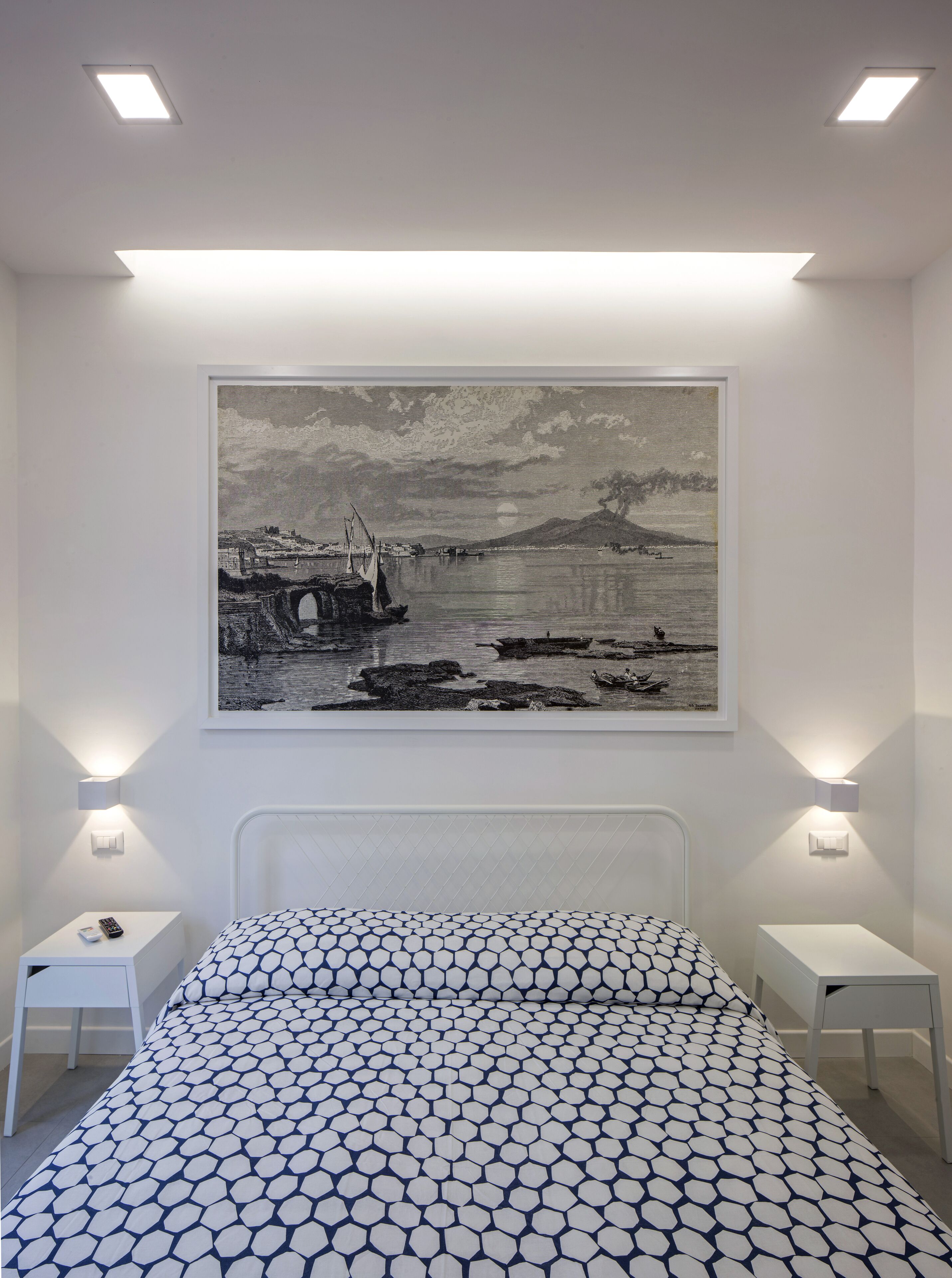 A bedroom with a bed, a nightstand, a wall-mounted painting, and two wall-mounted light fixtures.