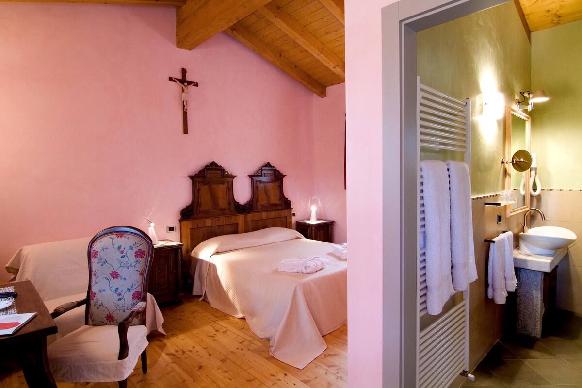 A bedroom with a wooden ceiling, a cross on the wall, a bed with a white bedspread, a chair with floral upholstery, and a bedside table with a lamp.