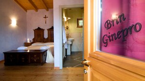 Una habitación de hotel con una cama de madera, una mesita de noche, una cruz en la pared y un baño visible a través de una puerta abierta.