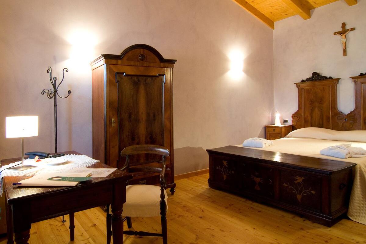 A bedroom with a wooden bed, a nightstand, a desk with a chair, a wardrobe, and a cross on the wall.