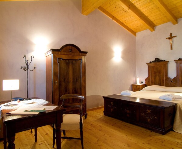 Un dormitorio con cama de madera, una mesita de noche, un escritorio con silla, un armario y una cruz en la pared.