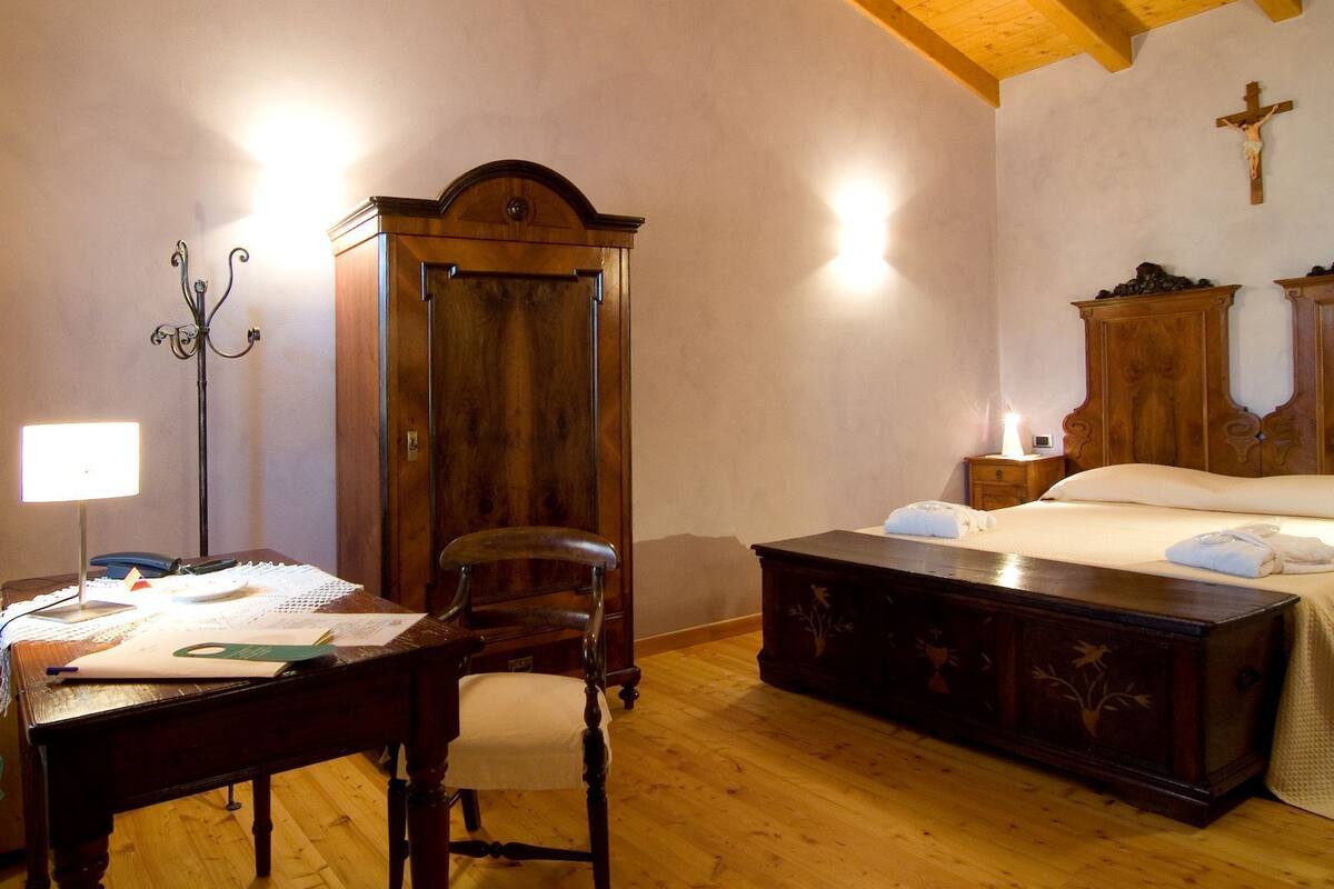 A bedroom with a wooden bed, a nightstand, a desk with a chair, a wardrobe, and a cross on the wall.