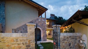 Casa de paredes de piedra con una escalera de piedra que lleva a una puerta, un pequeño jardín con flores rosas y un baño exterior visible.