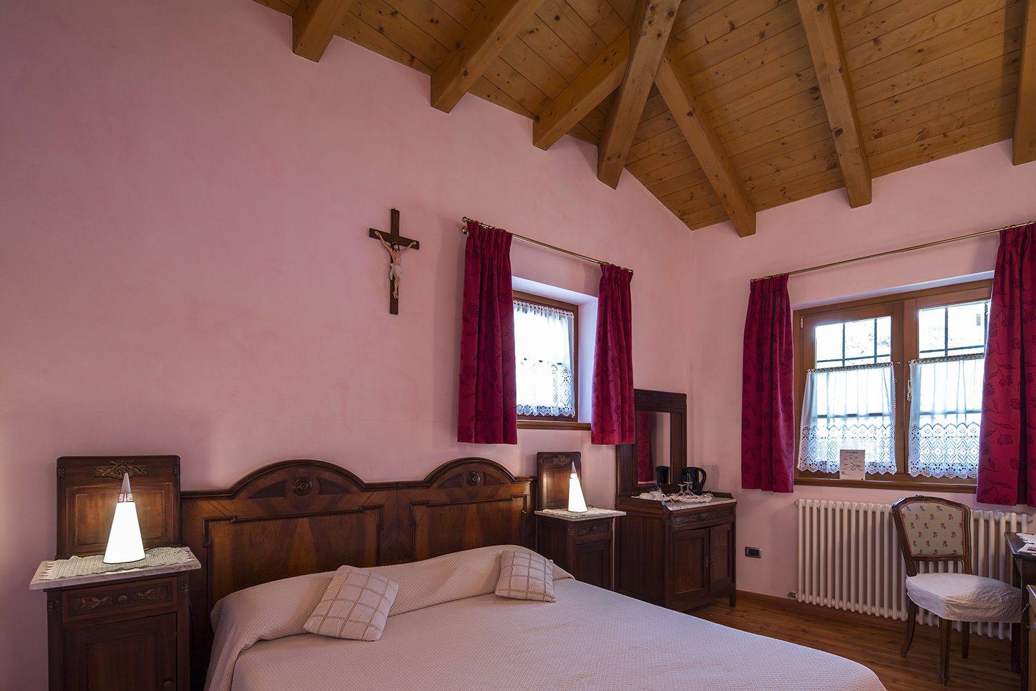A bedroom with a wooden ceiling, a cross on the wall, a bed with pillows, a nightstand with a lamp, a dresser, a mirror, and a chair.