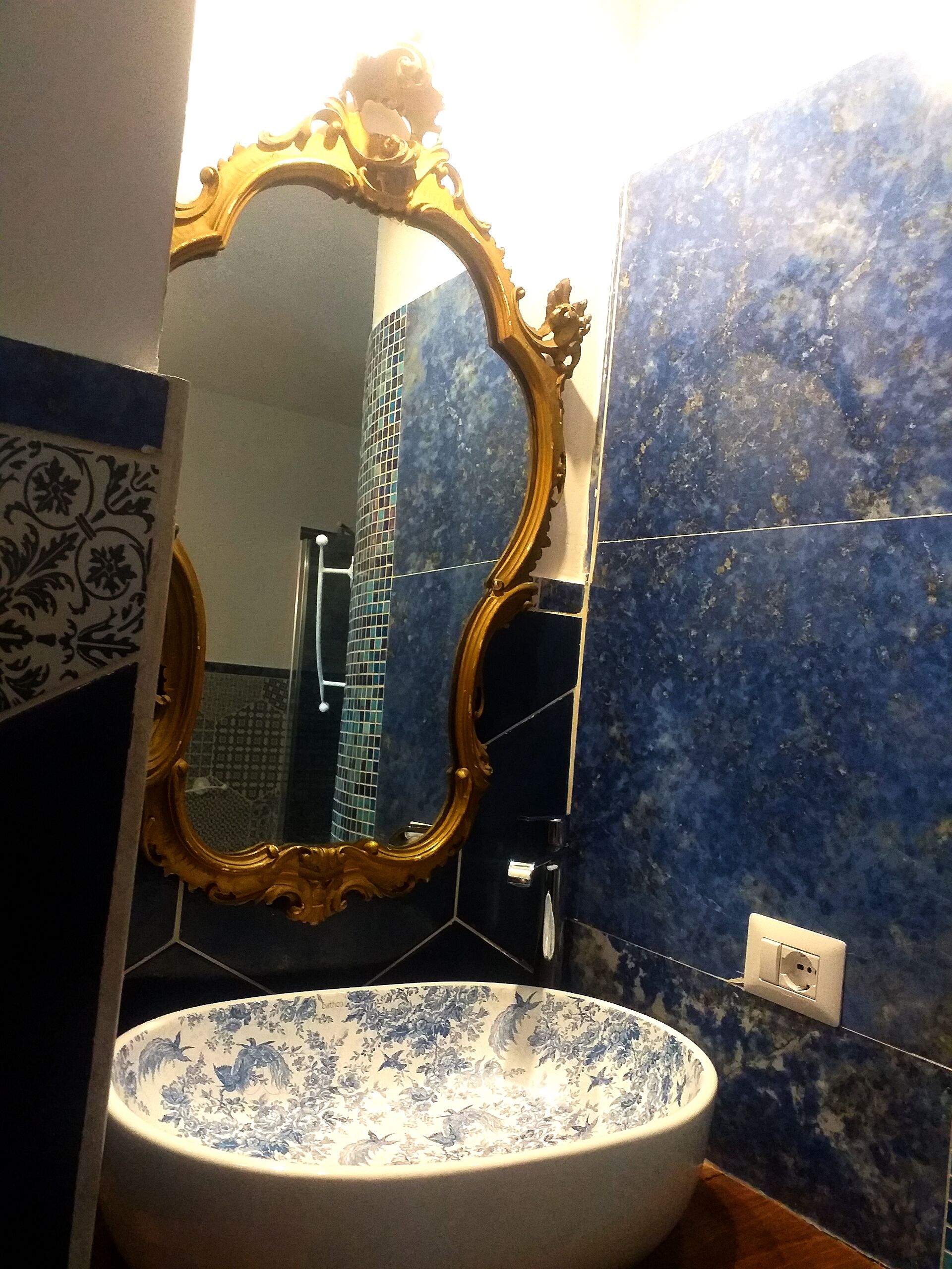 A bathroom with a large ornate mirror, a round sink with floral patterns, and blue marble tiles.