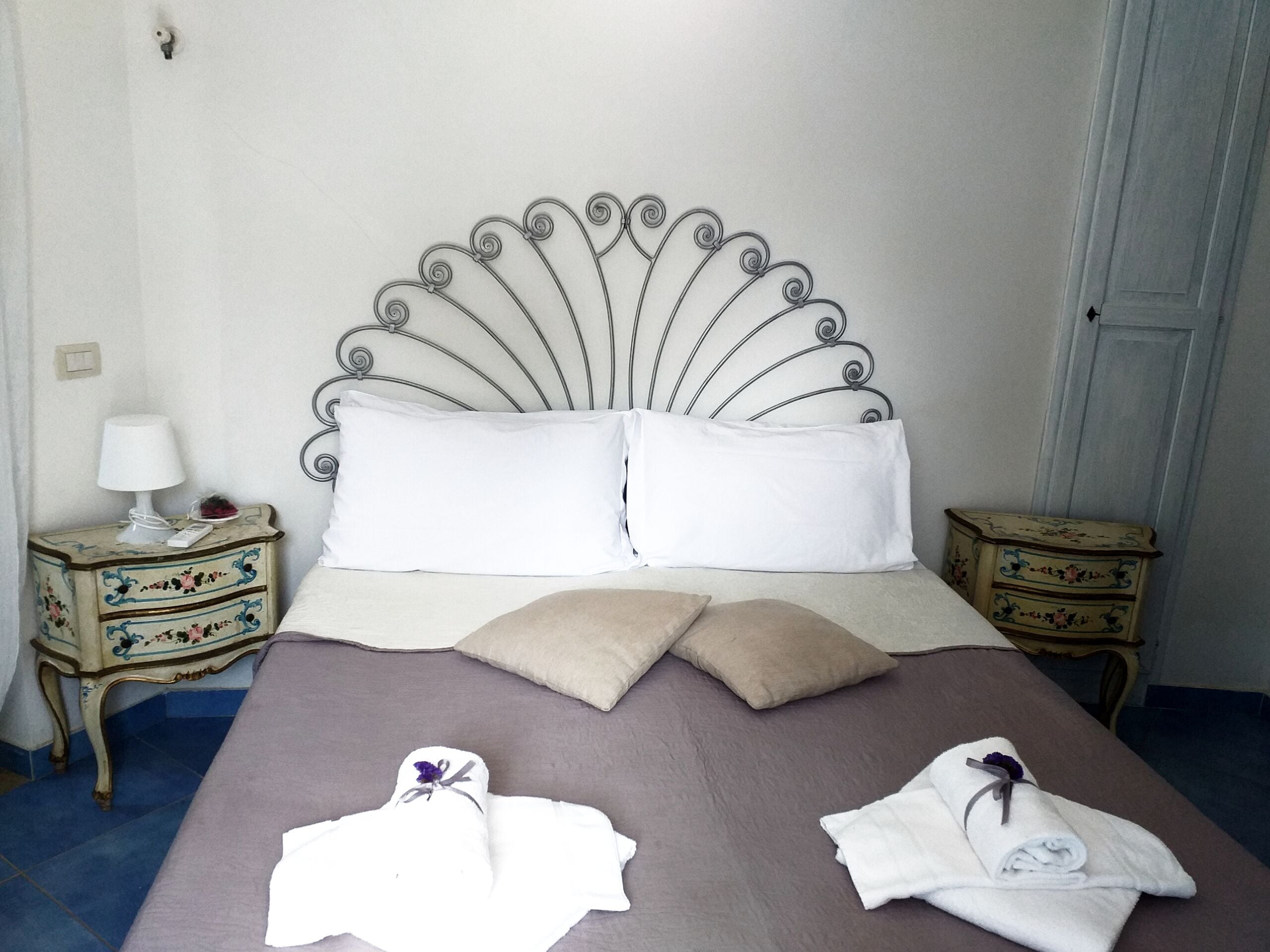 Una cama con un cabecero metálico decorativo, dos almohadas blancas, un edredón color beige y dos toallas sobre la cama.