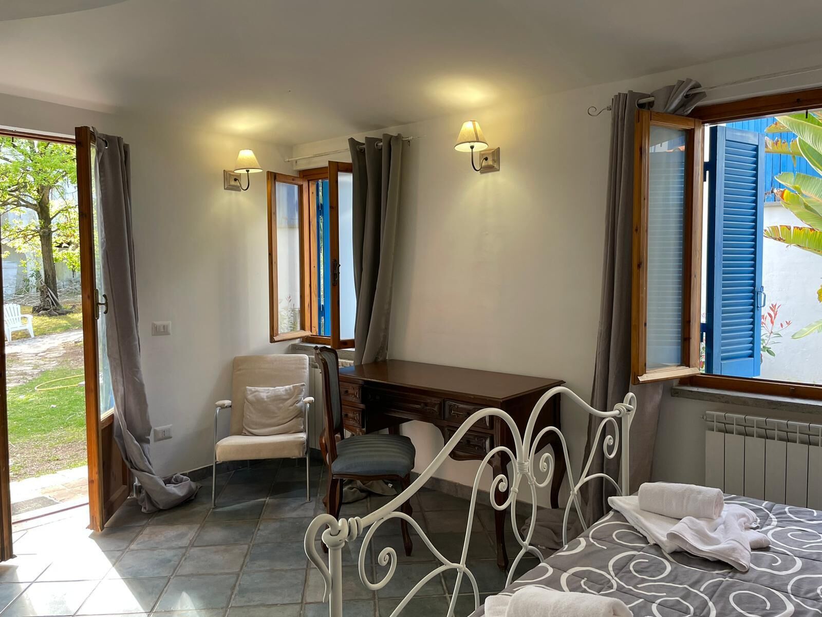 A bedroom with a bed, a desk, and a chair. There are two windows with curtains, one of which has a view of greenery outside.