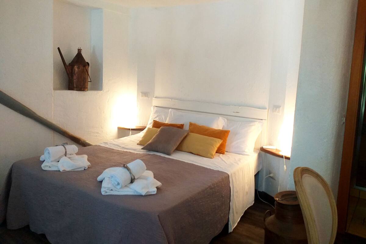 A hotel room with a bed, pillows, a nightstand, a chair, and a wall-mounted heater.
