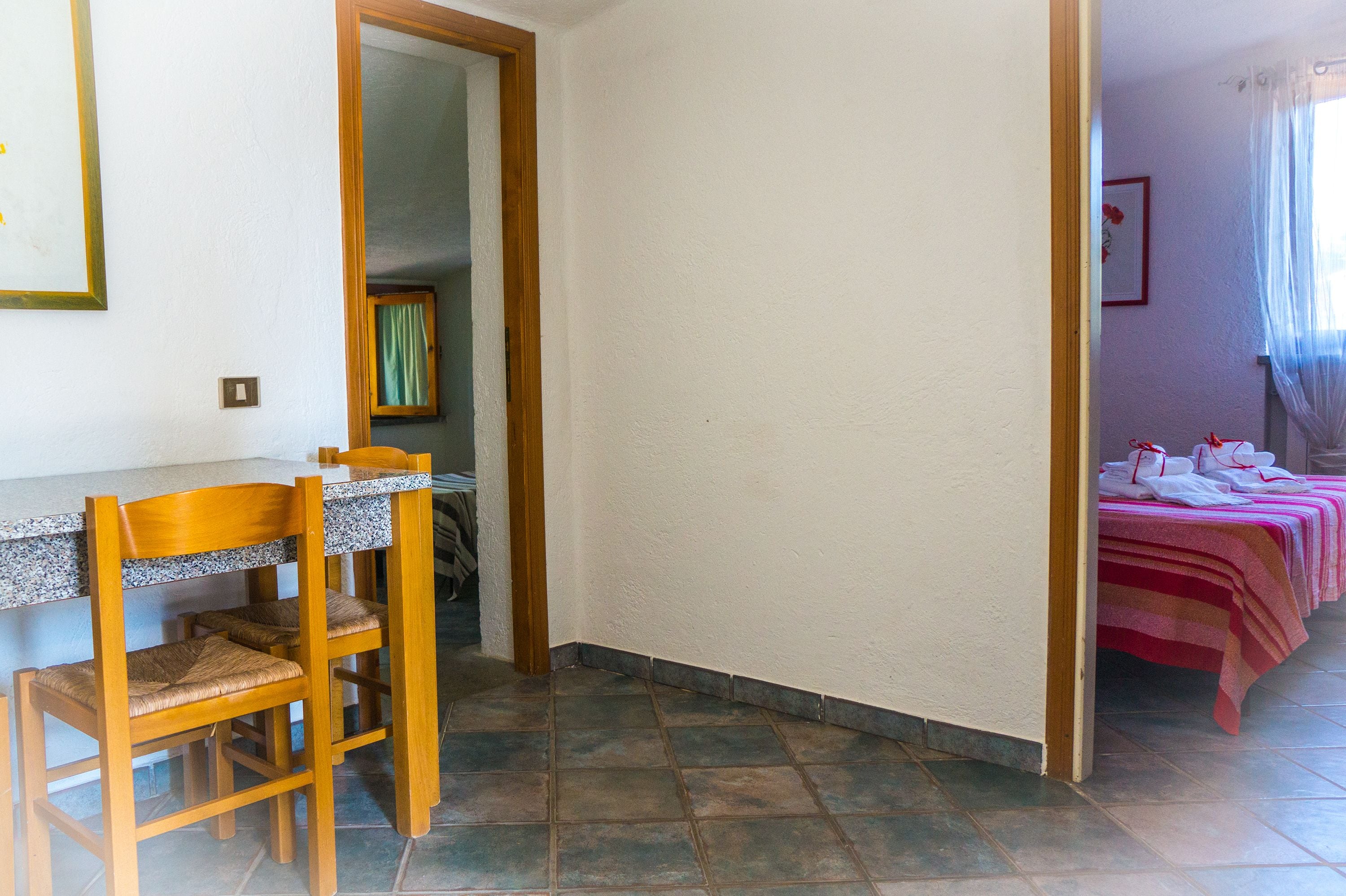 A room with a dining area featuring a wooden table and chairs, a bed with striped bedding, and a window with sheer curtains.