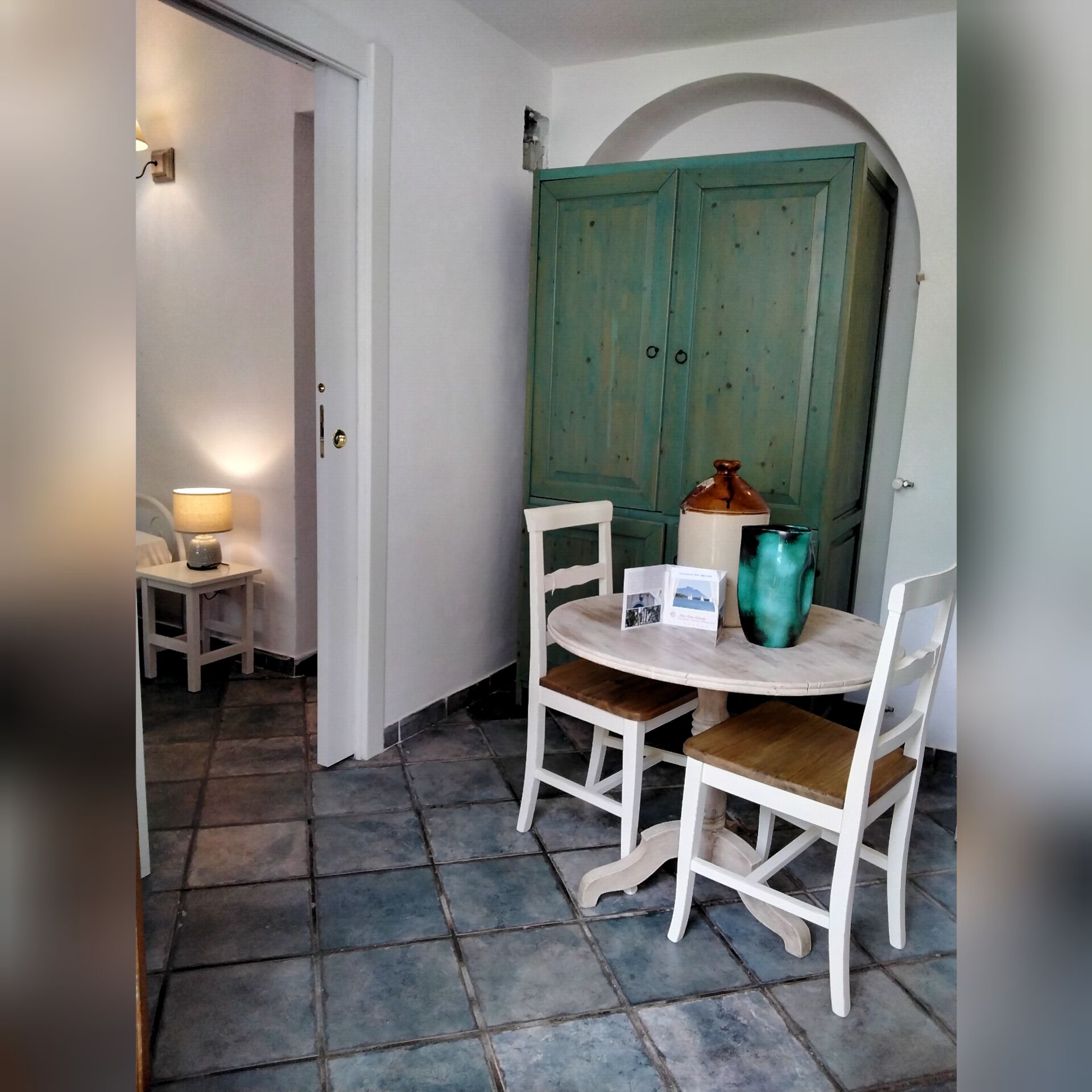 A small dining area with a table and chairs, a lamp, and a green door in the background.