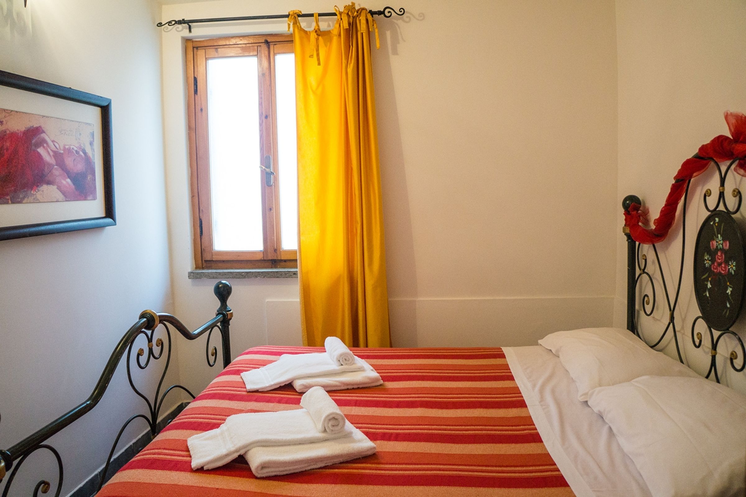 A hotel room with a bed, a window with yellow curtains, a framed picture on the wall, and a metal headboard with a red decoration.
