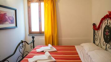 A hotel room with a bed, a window with yellow curtains, a framed picture on the wall, and a metal headboard with a red decoration.