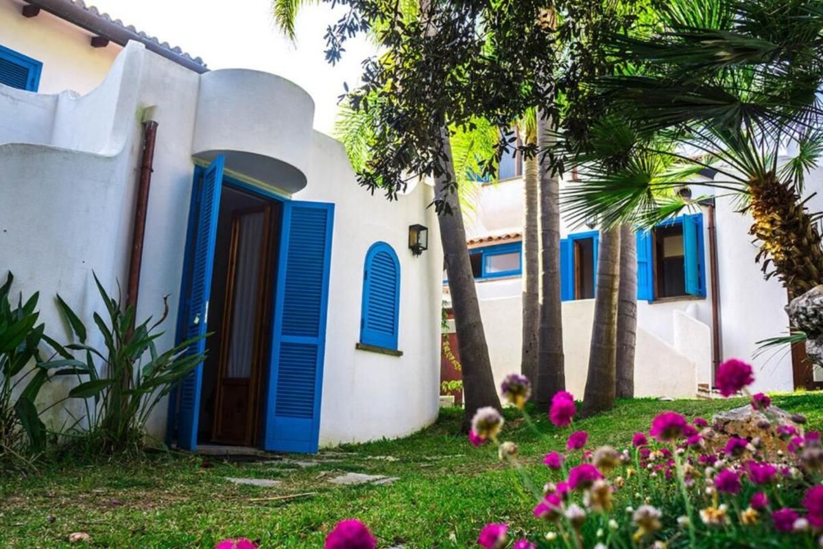 Edificio blanco con contraventanas azules y una puerta, rodeado de follaje y flores.