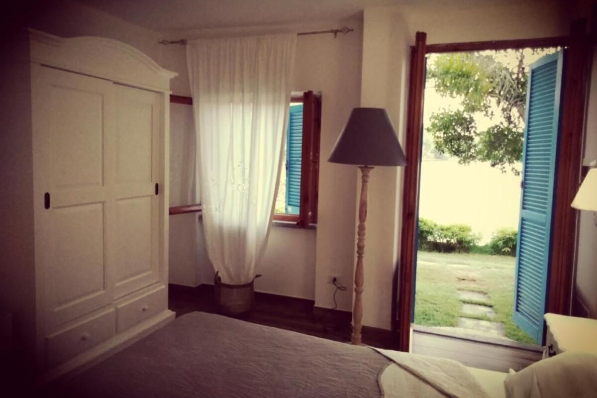 A bedroom with a bed, a window with blue shutters, a lamp, and a view of the outside.