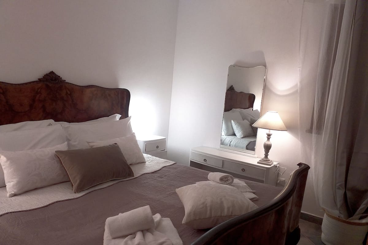 A neatly made bed with pillows and a blanket, a bedside table with a lamp, a mirror, and a wooden headboard.
