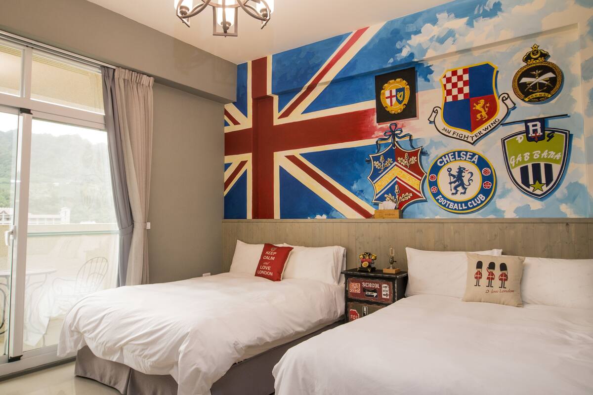 Habitación de hotel con dos camas, un ventanal grande con cortinas y una pared decorada con varios logotipos de clubes de fútbol.