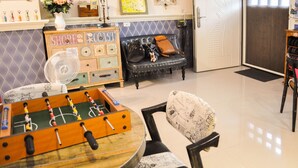 Una habitación con una mesa de futbolín, un sofá, una silla, un ventilador y una pared decorativa con un estante.