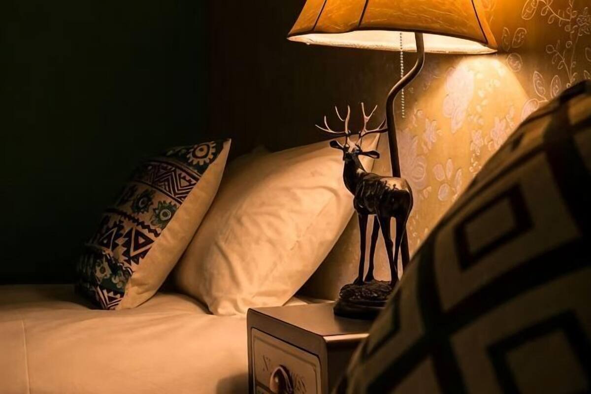 Una cama con almohadones estampados, una mesita de noche con una lámpara y una figurita de ciervo, y una pared con un diseño geométrico.