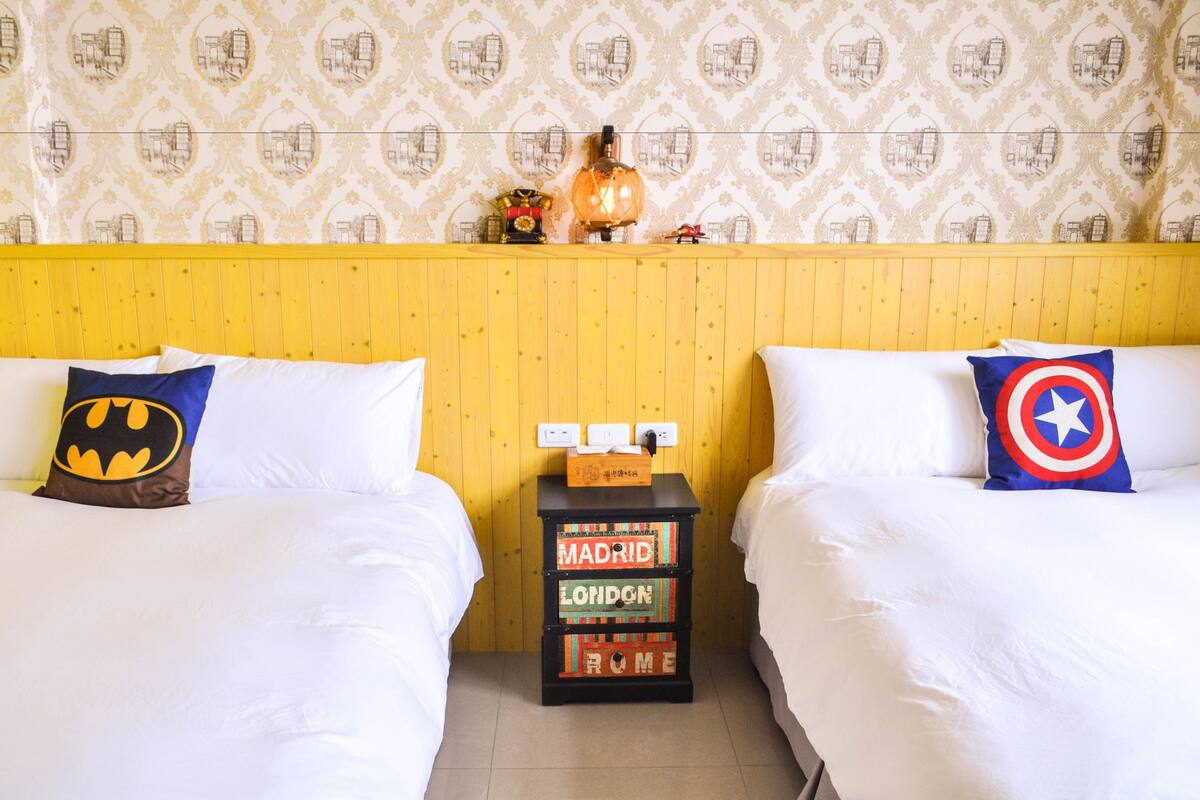 Habitación de hotel con dos camas, cada una con un cojín temático, un cabecero de madera y una mesita de noche con una lámpara y una caja.