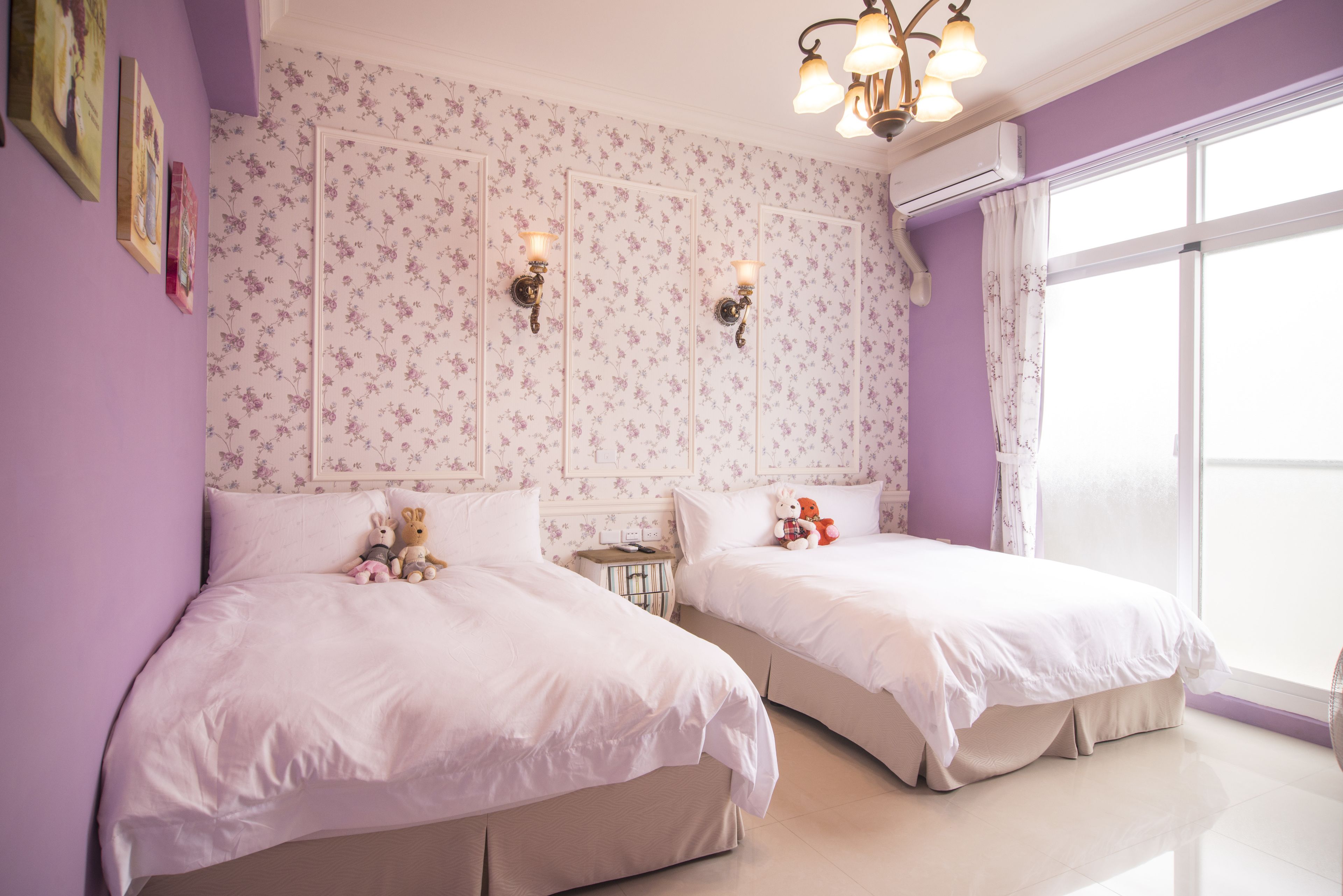 A bedroom with two beds, floral wallpaper, wall-mounted lights, and a large window.