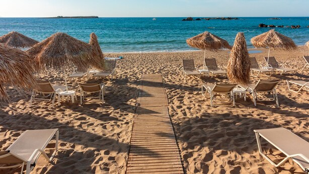 Una playa arenosa con un camino de madera que lleva a sillas de playa y sombrillas de paja.