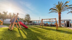 Un parque infantil con un tobogán y columpios, una palmera y una vista de una playa y sombrillas al fondo.