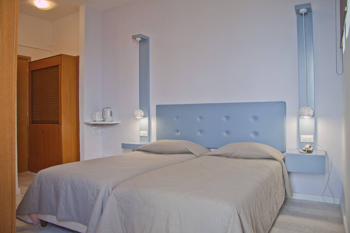 A bedroom with two beds, a wooden door, a nightstand with a lamp, and a shelf with a kettle.