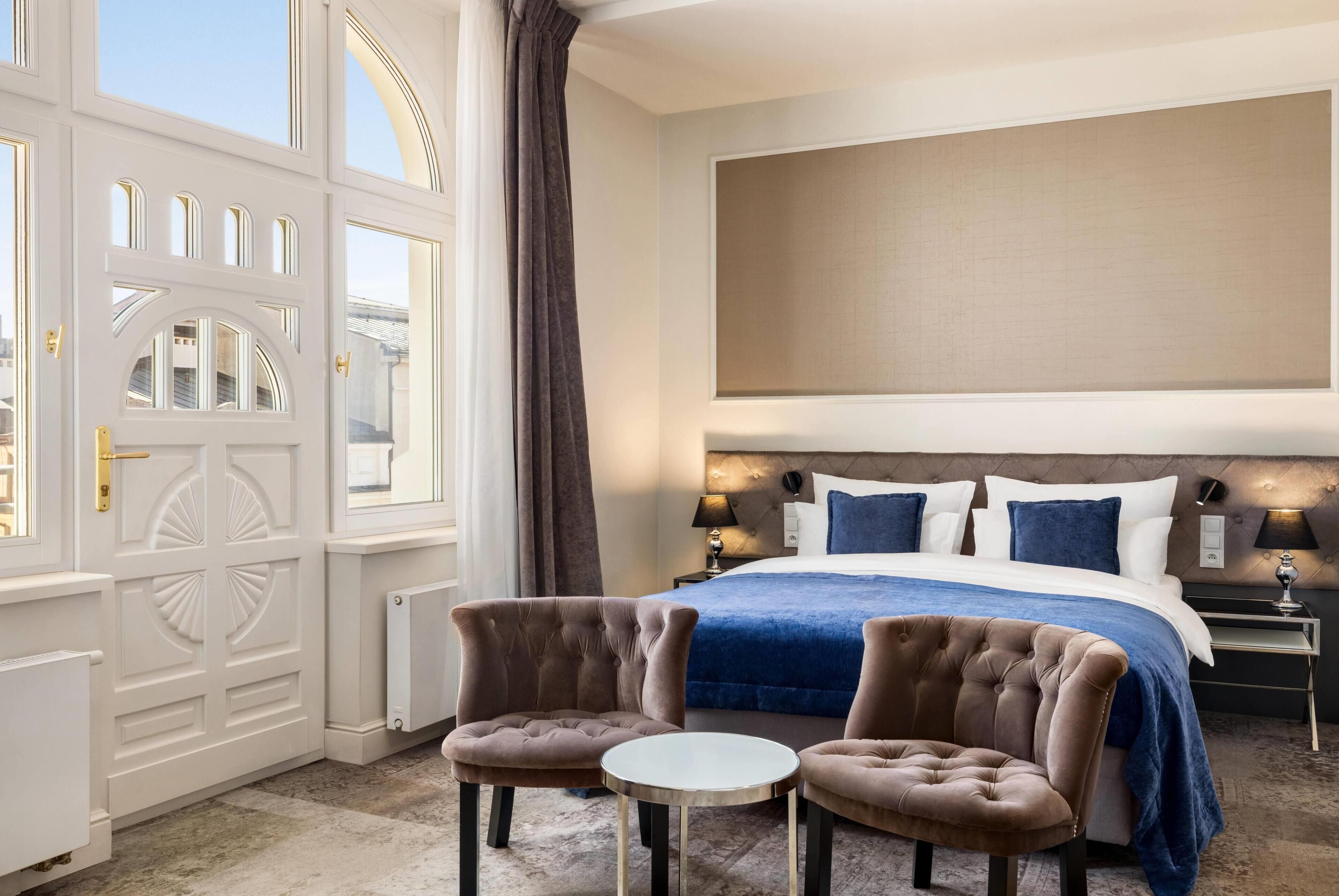 A hotel room with a large bed, two armchairs, a small table, a large window with curtains, and a view of the city.