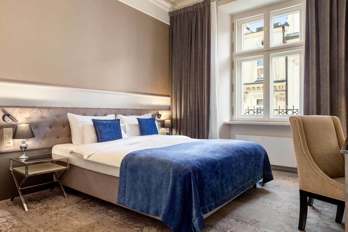Hotellihuone, jossa on suuri sänky, yöpöytä, tuoli, ikkuna, josta on näkymä rakennuksiin, ja lamppu.