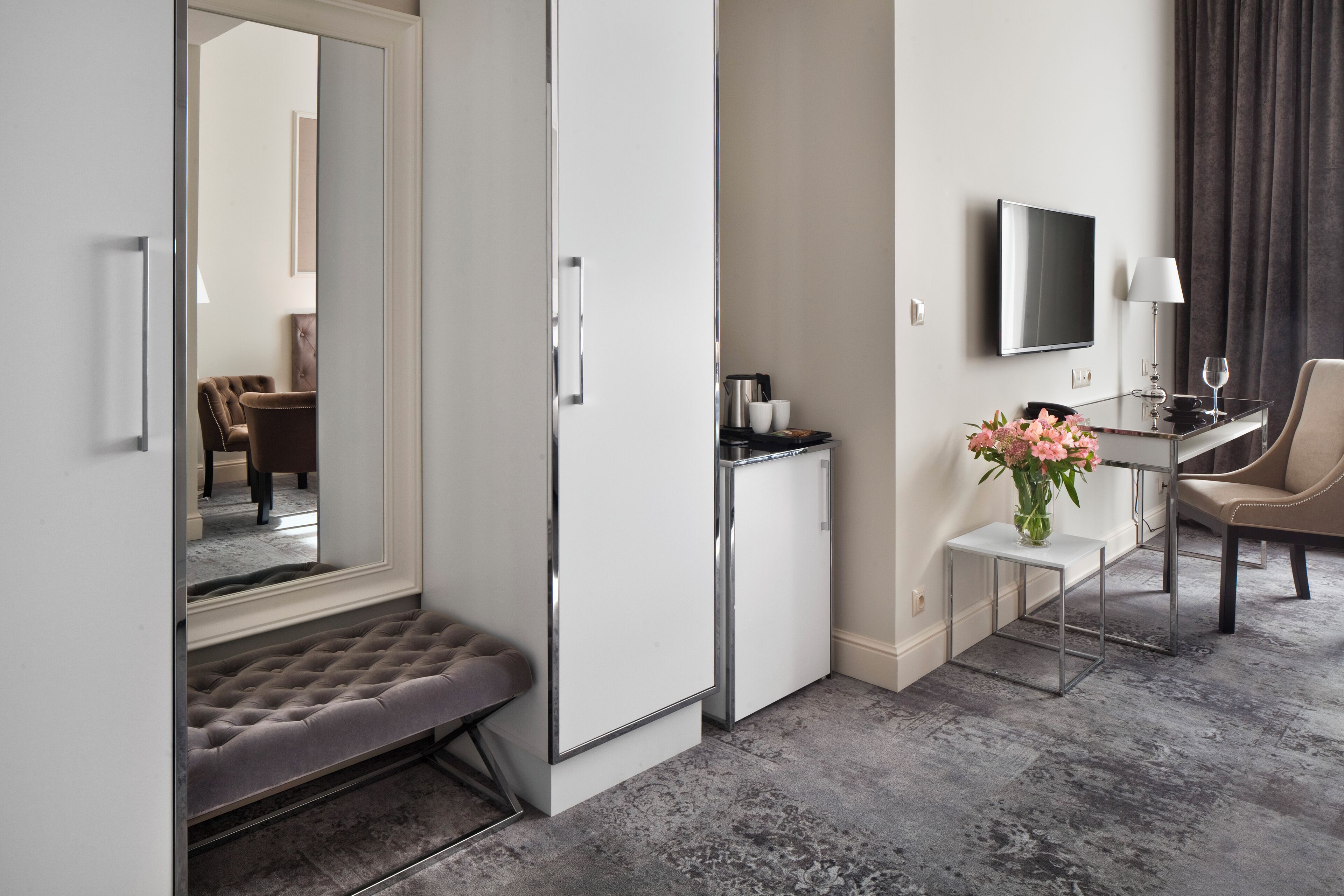 Una habitación de hotel con una cama, una mesita con un florero, un televisor y un rincón de descanso con una silla.